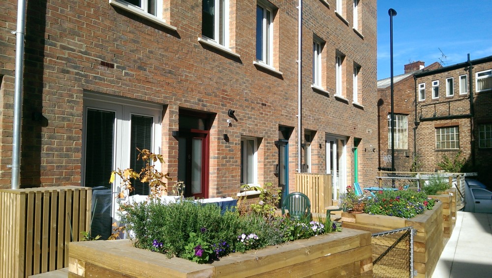 Three homes with brightly coloured front doors are fronted by small gardens with low, wooden planters separating them